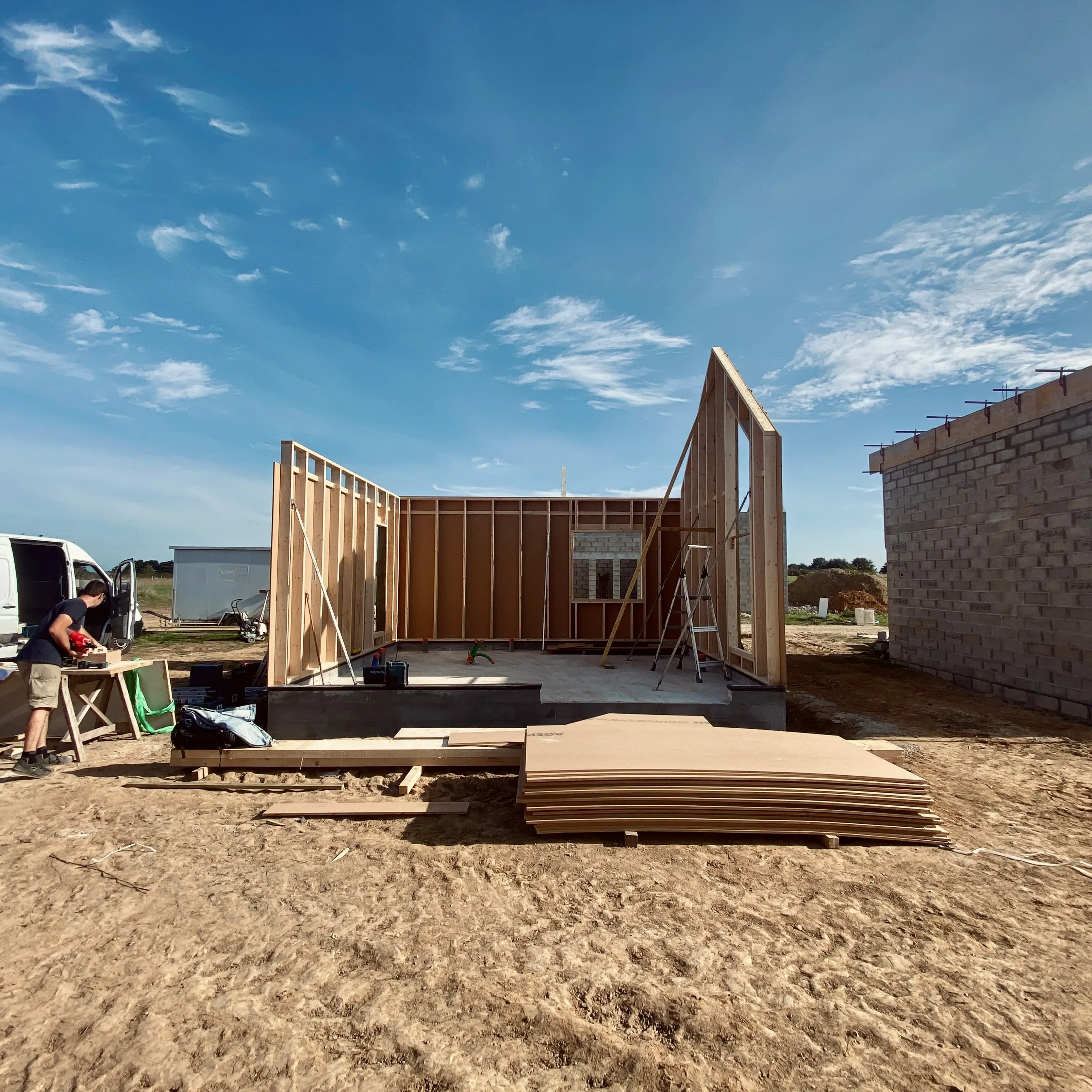 Chantier d’une maison en ossature bois : murs dressés sur la dalle, panneaux de bois empilés au premier plan et un compagnon au travail